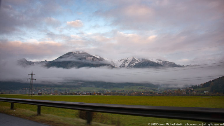 Snow-dusted mountain range beyond a foggy Alpine valley, with fields, settlement, power lines, and highway guardrail.