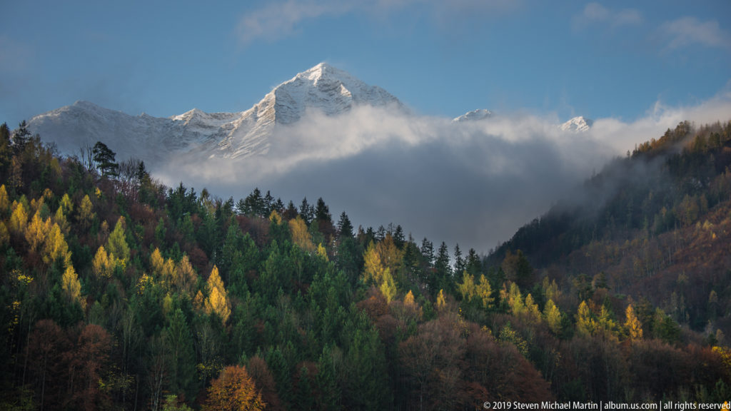 Snowy Alpine summit above low clouds and mixed autumn forest with yellow larches and dark conifers.