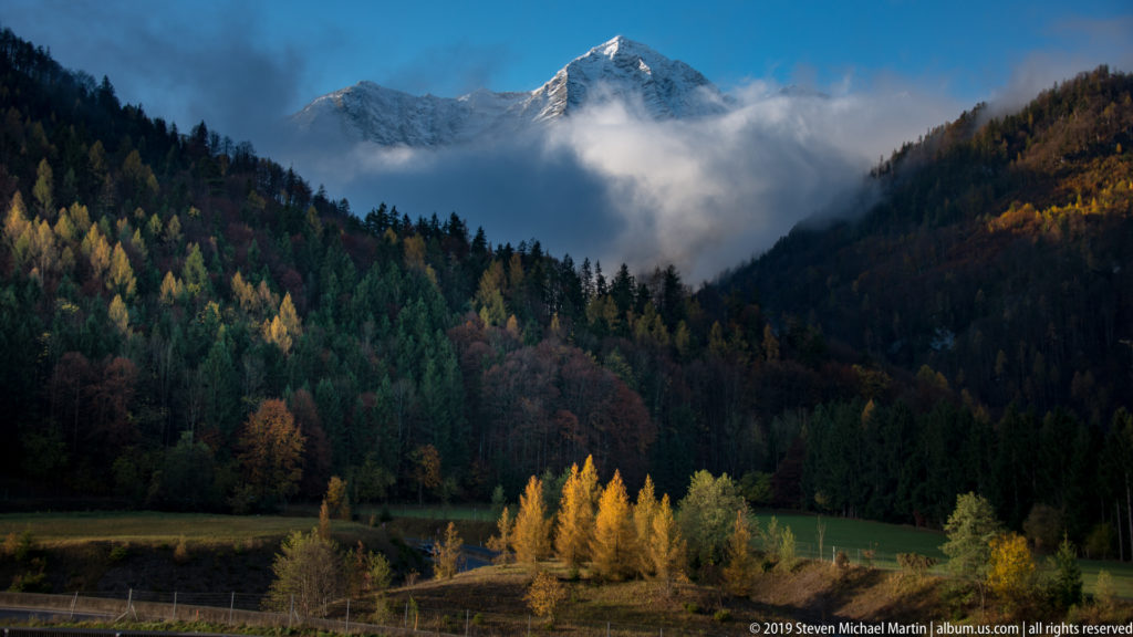 Autumn forest and green field below a snow-covered Alpine peak partly hidden by low cloud.