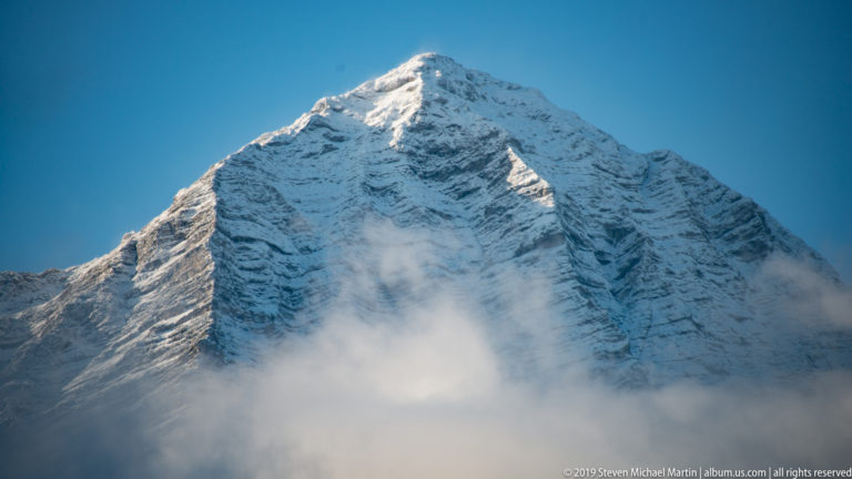 Steep snow-covered pyramidal Alpine peak rising above a low bank of cloud under blue sky.