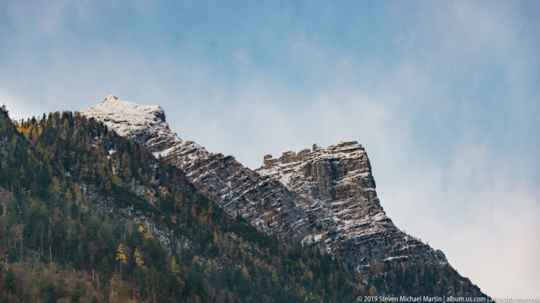 Jagged stratified mountain ridge with snow on upper ledges and autumn forest on the lower slope.
