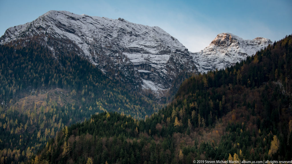 Snow-covered craggy Alpine peaks rising above dark conifer forest in autumn.