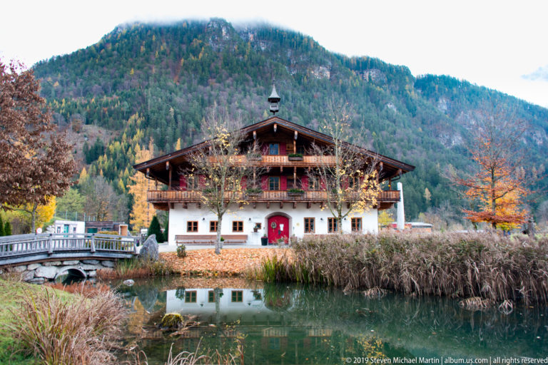 Timber chalet with balconies and red doorway beside a reflective pond, with a wooded mountainside behind.