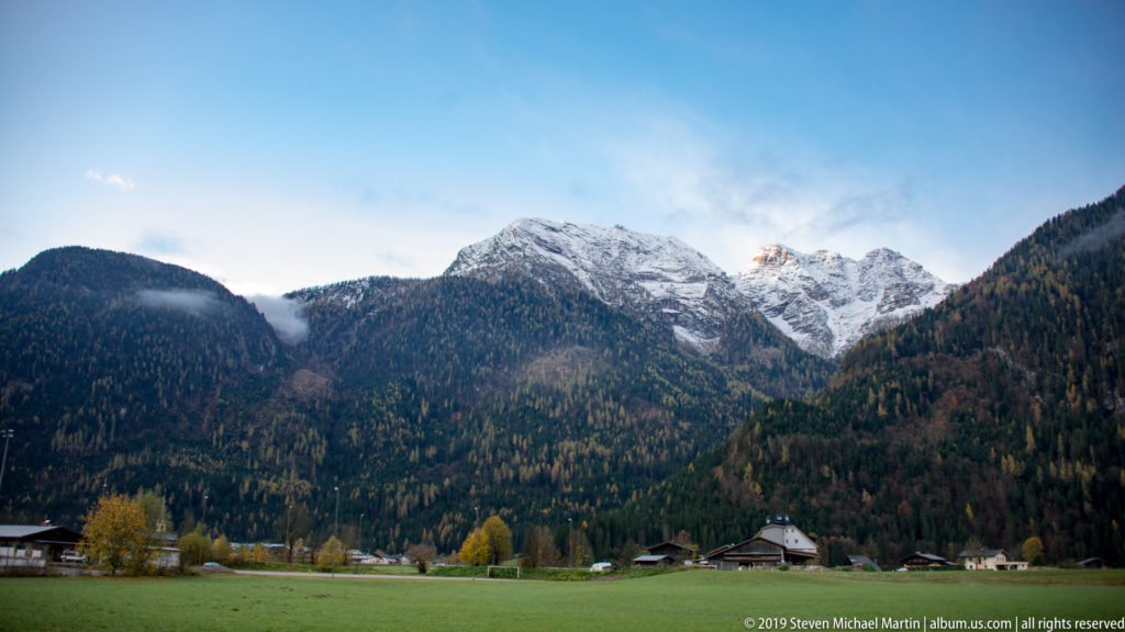 Green valley meadow and scattered houses below forested slopes and snow-dusted Austrian Alpine peaks.