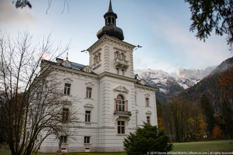 White multi-story residence with a square tower and dark roof, backed by snow-covered Alpine peaks and autumn trees.