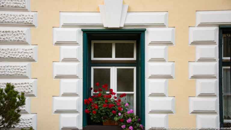 Dark-framed window with red and pink geraniums, surrounded by white stucco blocks on a pale yellow façade.