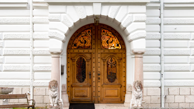 Arched carved wooden double doors with black iron grilles, flanking columns, and small lion sculptures.