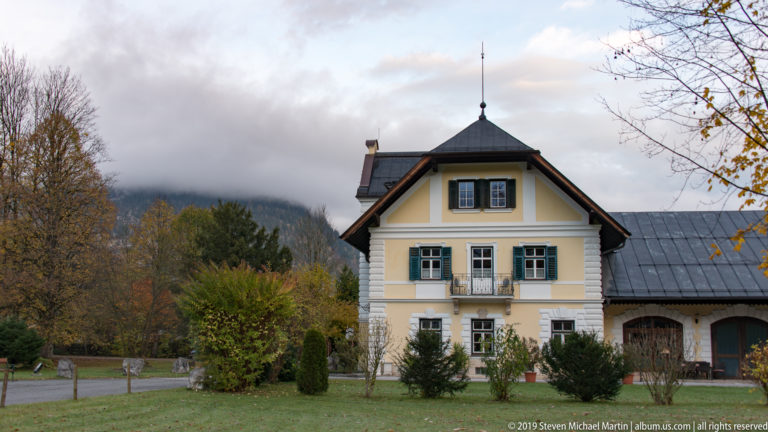 Pale yellow Alpine house with green shutters and balcony attached to a long outbuilding beneath a cloudy hillside.