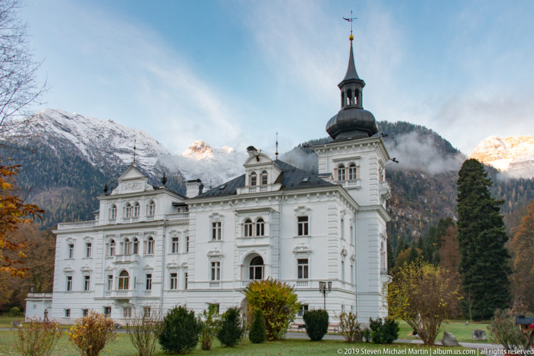 Ornate white multi-story building with central cupola and shaped gables against snowy Alpine mountains.