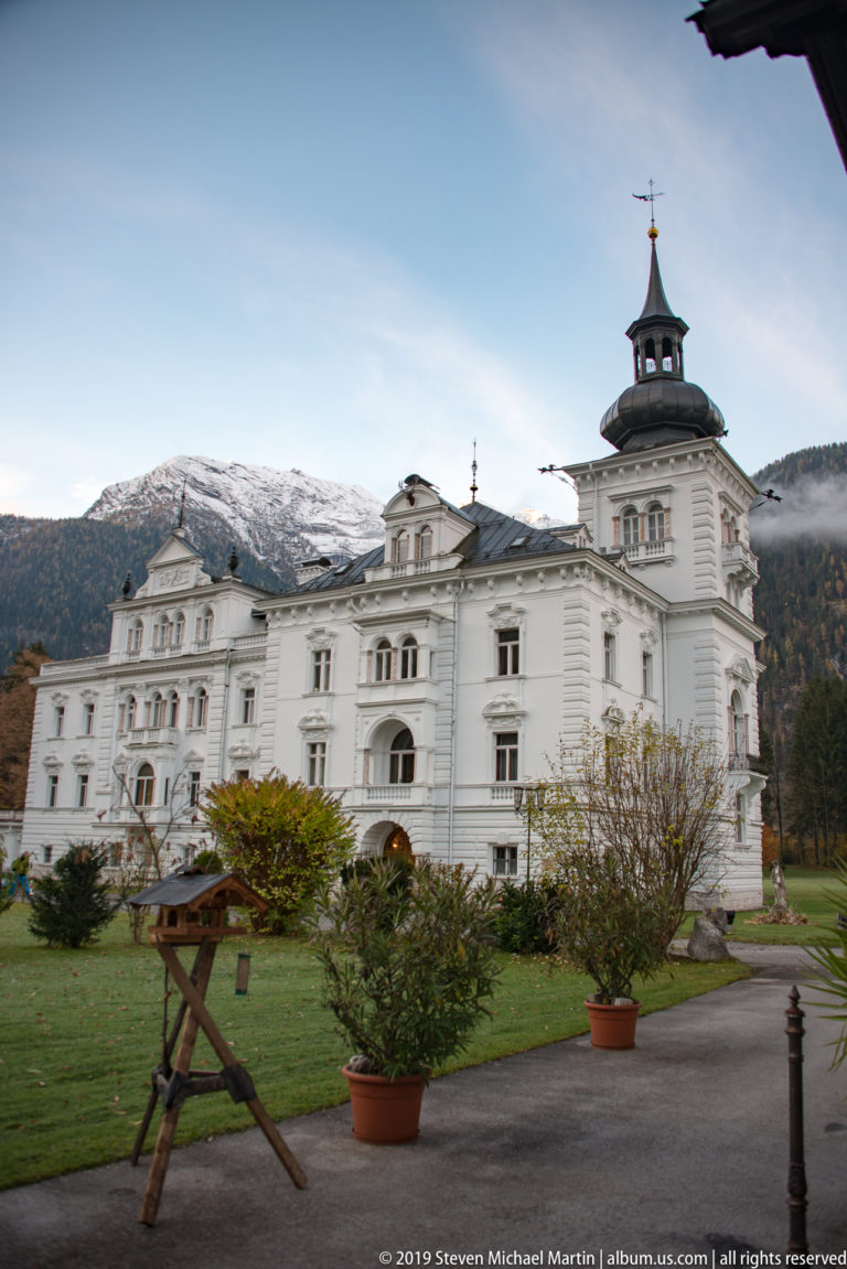 Large ornate white building with central cupola tower in landscaped grounds, backed by a snow-dusted mountain ridge.