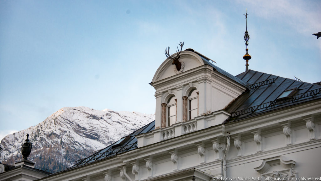 Ornate white dormer and cornice with antlers at the gable crest, backed by a snow-covered mountain.
