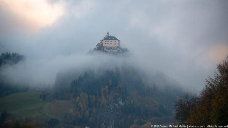 Pale hilltop building with dark roof above thick fog, with autumn forest and green slopes partly visible below.