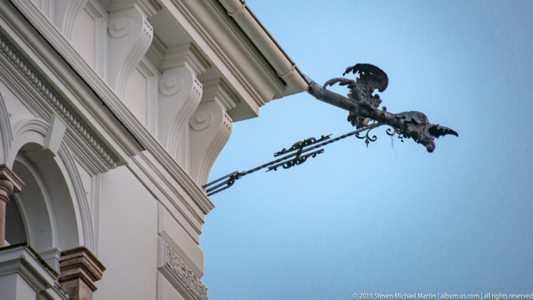 Ornate projecting metal bracket with a winged dragon-like figure attached to a white historic façade.