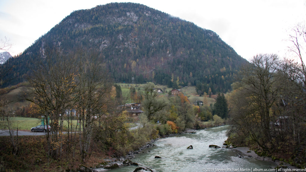 Rocky river flowing through an autumn valley with trees, scattered houses, pasture, and a wooded mountain.
