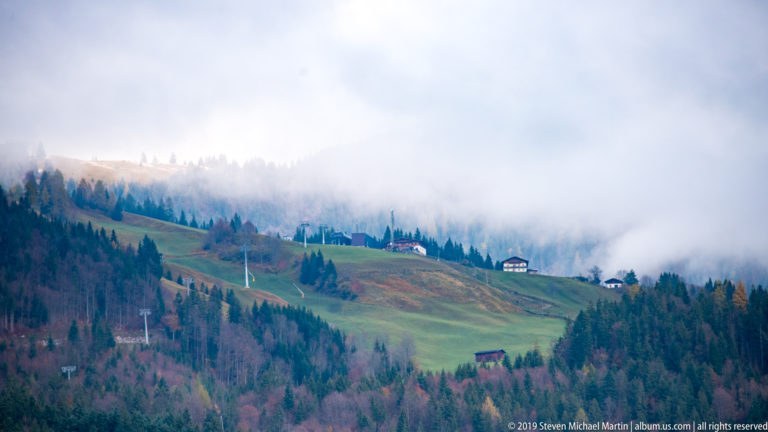 Green Alpine hillside with scattered buildings and conifers partly swallowed by dense low cloud.