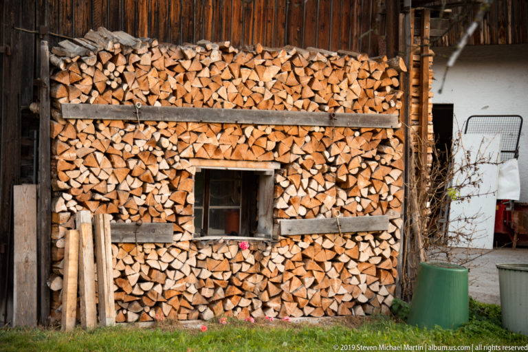 Dense stack of split firewood arranged around a small window beneath a dark timber shed wall.