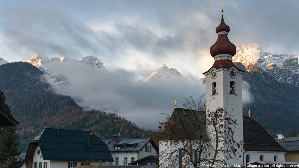 White onion-domed church tower with clock faces beneath misty, snow-streaked mountains.