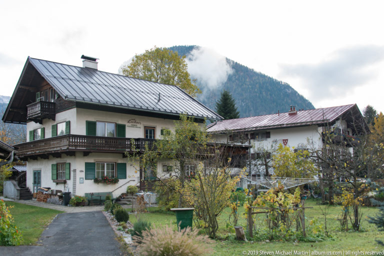 White farmhouse with green shutters and balconies beside a garden, outbuildings, and misty wooded mountain.