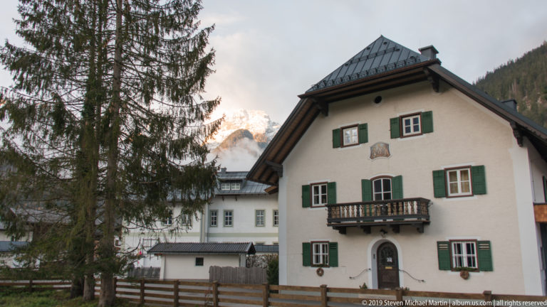 White Alpine house with green shutters, dark central balcony, arched doorway, and small painted emblem near the roof.