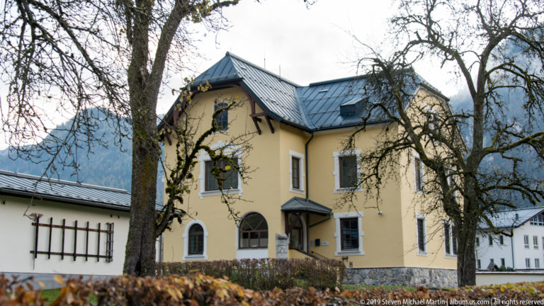 Pale yellow villa with steep gray metal roof, projecting gable, arched windows, and decorative corner trim.