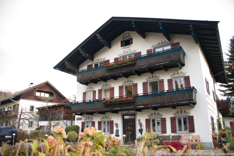 White multi-story Alpine house with dark wooden balconies, red shutters, flower boxes, and painted window decoration.