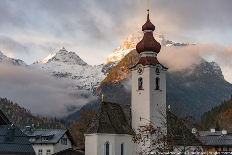 White church tower with red-brown onion dome and clocks in front of sunlit snowy mountains and low cloud.