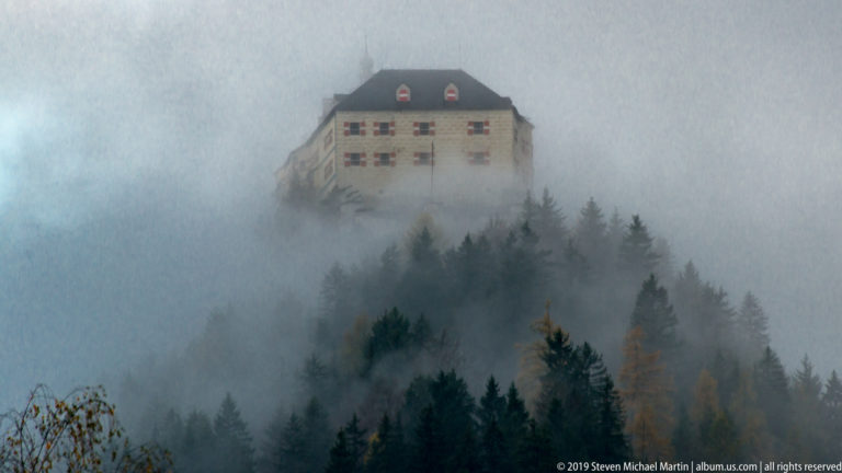 Large pale hilltop building with dark roof and dormers emerging through dense fog above conifer and autumn forest.