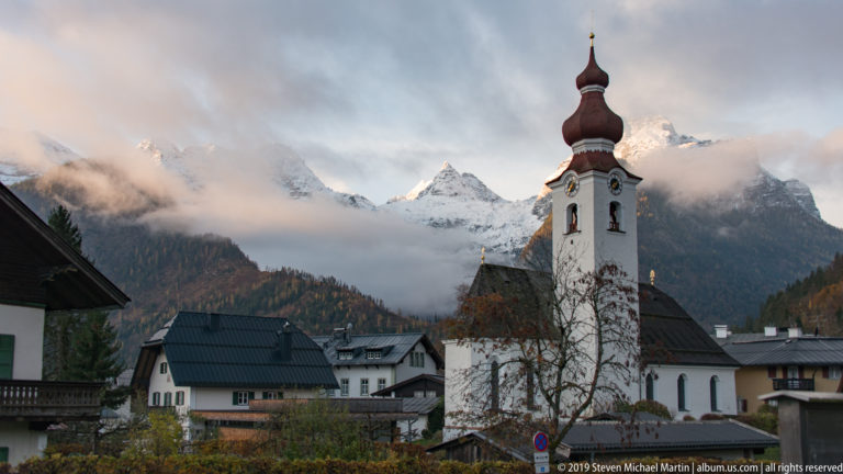 White onion-domed church tower among village houses, backed by cloud-wrapped snowy Alpine peaks.