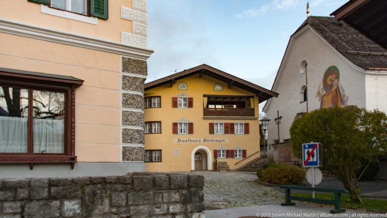 Yellow gasthaus with shutters and balcony beside a small white church bearing a painted religious mural.