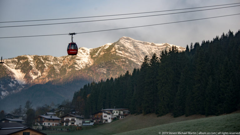 Red gondola suspended on a cable above village houses, with forest and a sunlit mountain behind.