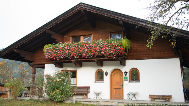 White Alpine house with dark timber gable, wooden balcony overflowing with red and pink flowers, and arched door.