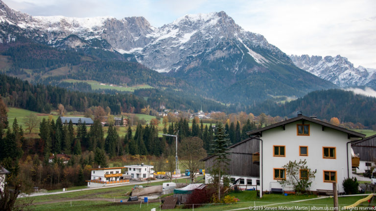 Houses and conifers in an Alpine valley below jagged mountains with fresh snow on upper slopes.