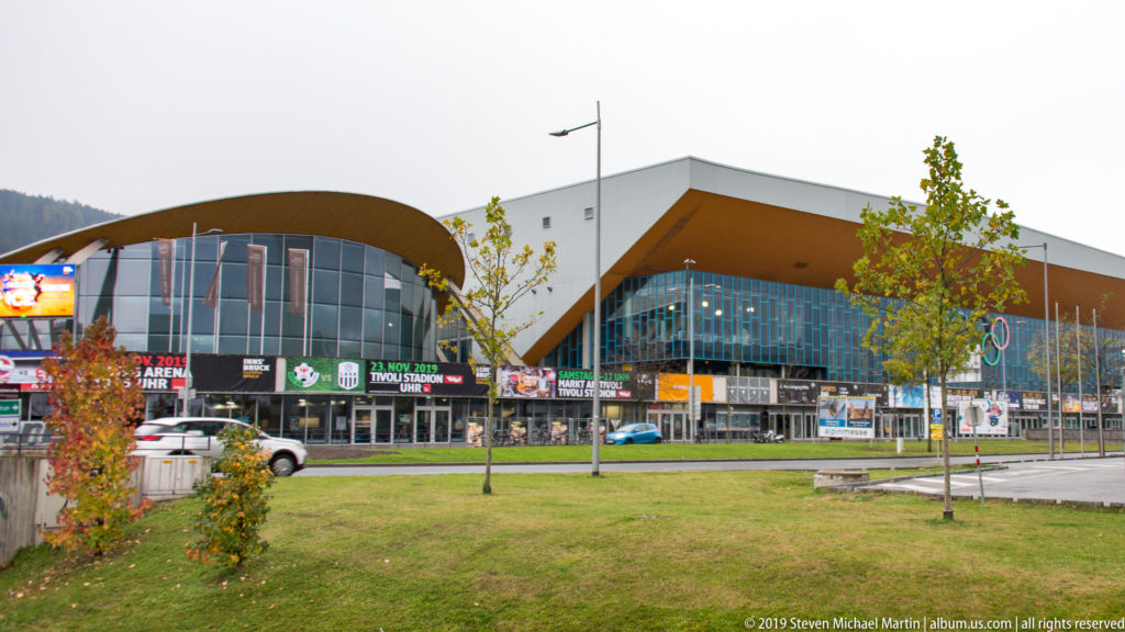 Large modern sports complex with curved roof and glass façades beyond a lawn and young trees.