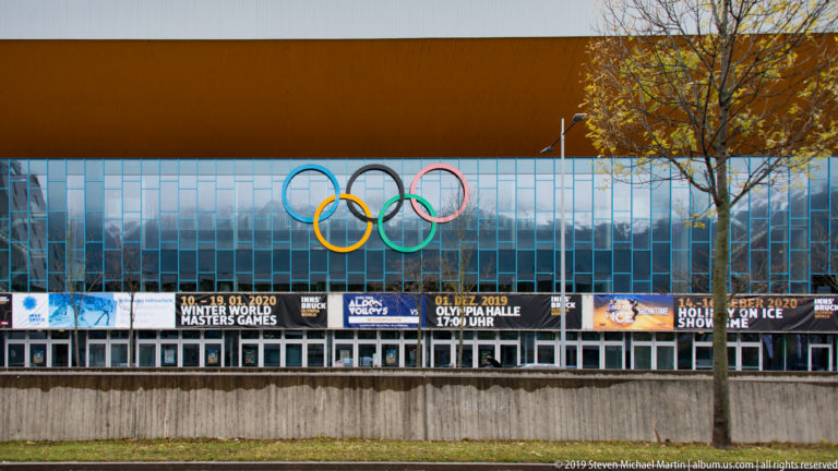 Olympic rings mounted on a glass sports-arena façade beneath a broad roof, with event banners along the entrance.