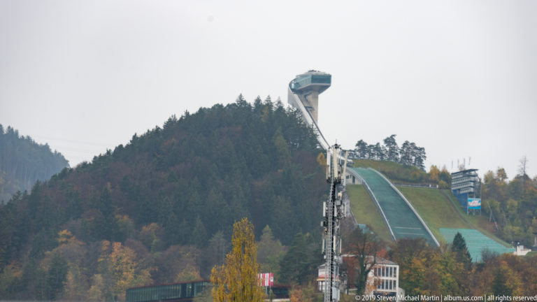 Distant Bergisel ski-jump tower and ramp rising above autumn trees and foreground structures.
