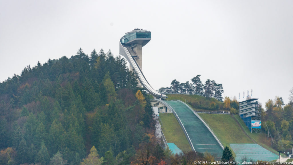 Bergisel Ski Jump tower and long descending ramp centered above a forested autumn hillside.