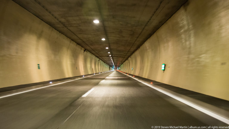 Straight roadway inside a concrete tunnel with ceiling lights, lane markings, and illuminated emergency indicators.