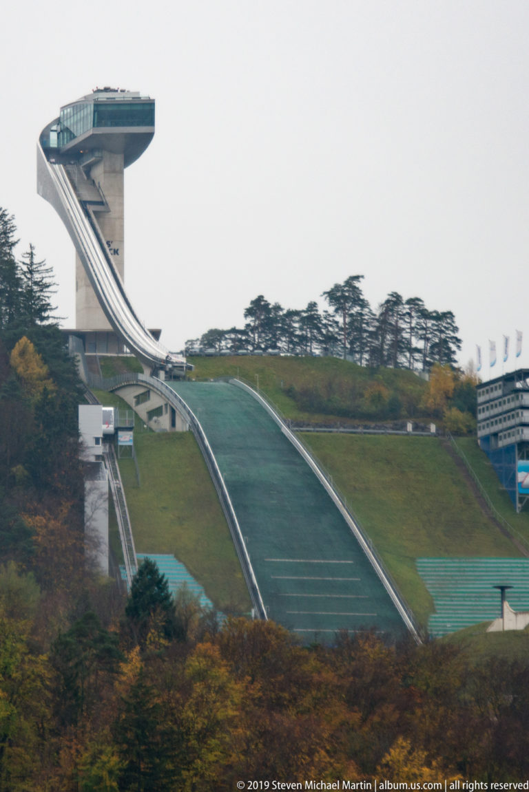 Green ski-jump landing hill with elevated ramp at left and support buildings beside a wooded slope.