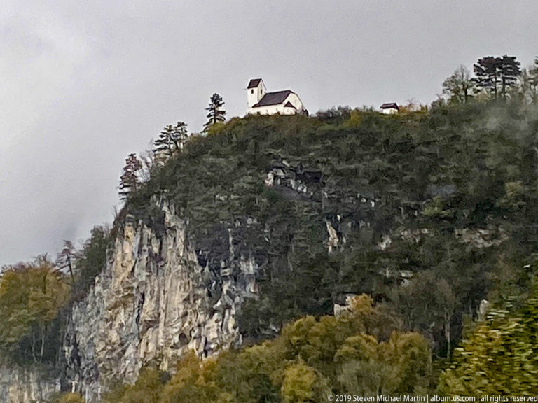 Small white chapel on the wooded crest of a steep pale limestone cliff in the Austrian Alps.
