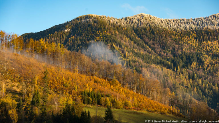 Sunlit Alpine slopes covered with golden larches and dark conifers, with a small patch of mist above meadow.
