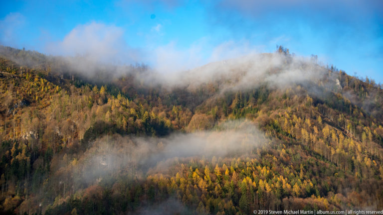 Bands of mist crossing a densely forested Alpine slope in yellow, orange, green, and brown autumn colors.