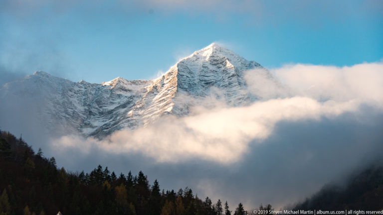 Sunlit snow-covered Alpine peak rising above dense white clouds, with dark forest along the lower edge.