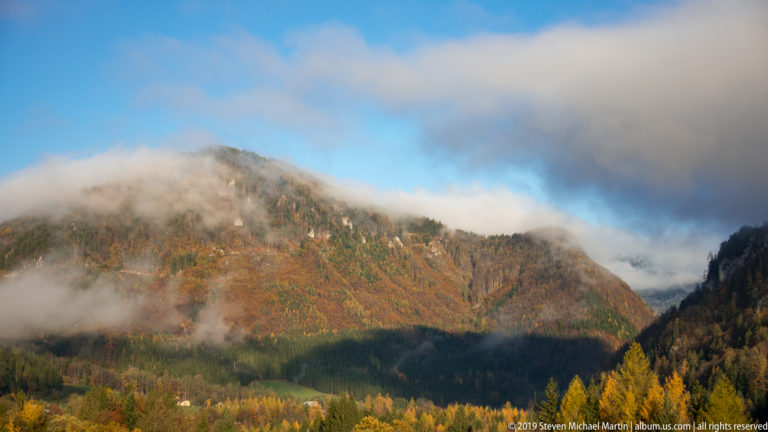 Low clouds crossing an autumn-colored Alpine mountainside beneath patches of blue sky.