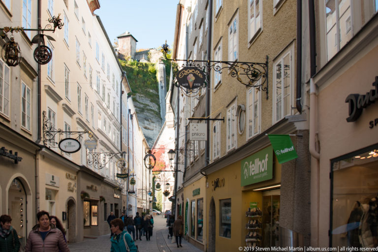 Pedestrians fill narrow Getreidegasse between historic shopfronts and wrought-iron signs, with the rocky Mönchsberg slope visible at the far end.