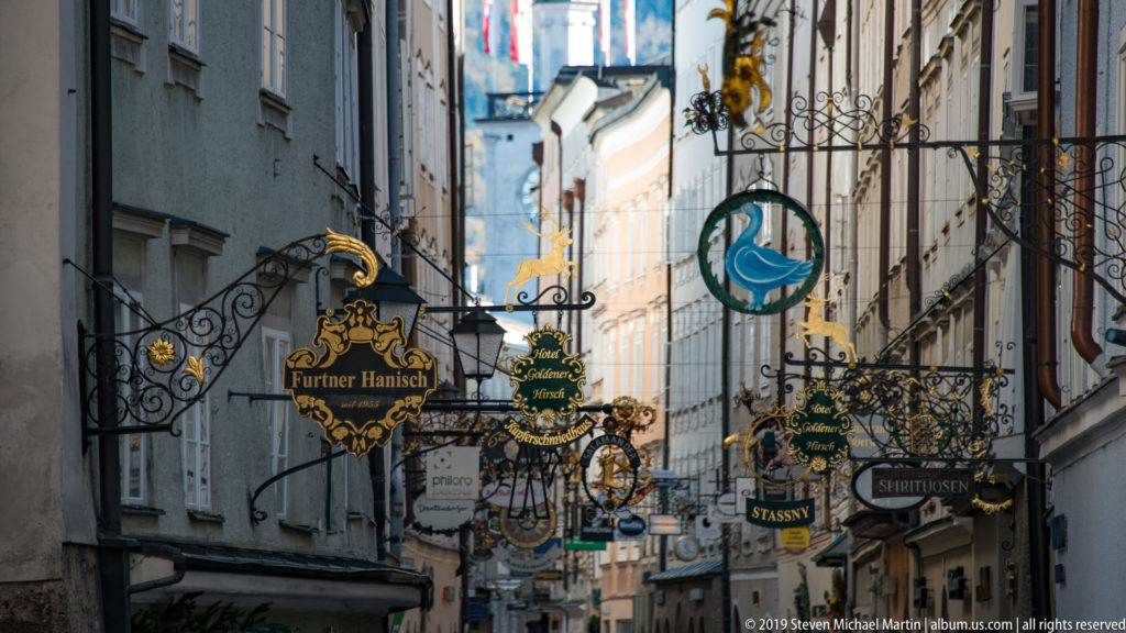 Dense cluster of ornate projecting shop signs spanning Getreidegasse, including Fürster Hansl and Stassny signs.