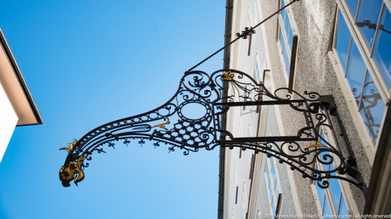 Ornate triangular wrought-iron sign bracket with scrollwork and a gilded animal-head finial against blue sky.