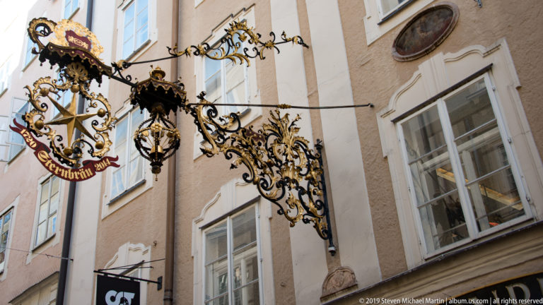 Large black-and-gold wrought-iron trade sign with dense scrollwork and suspended emblems projecting from a pale stucco façade.