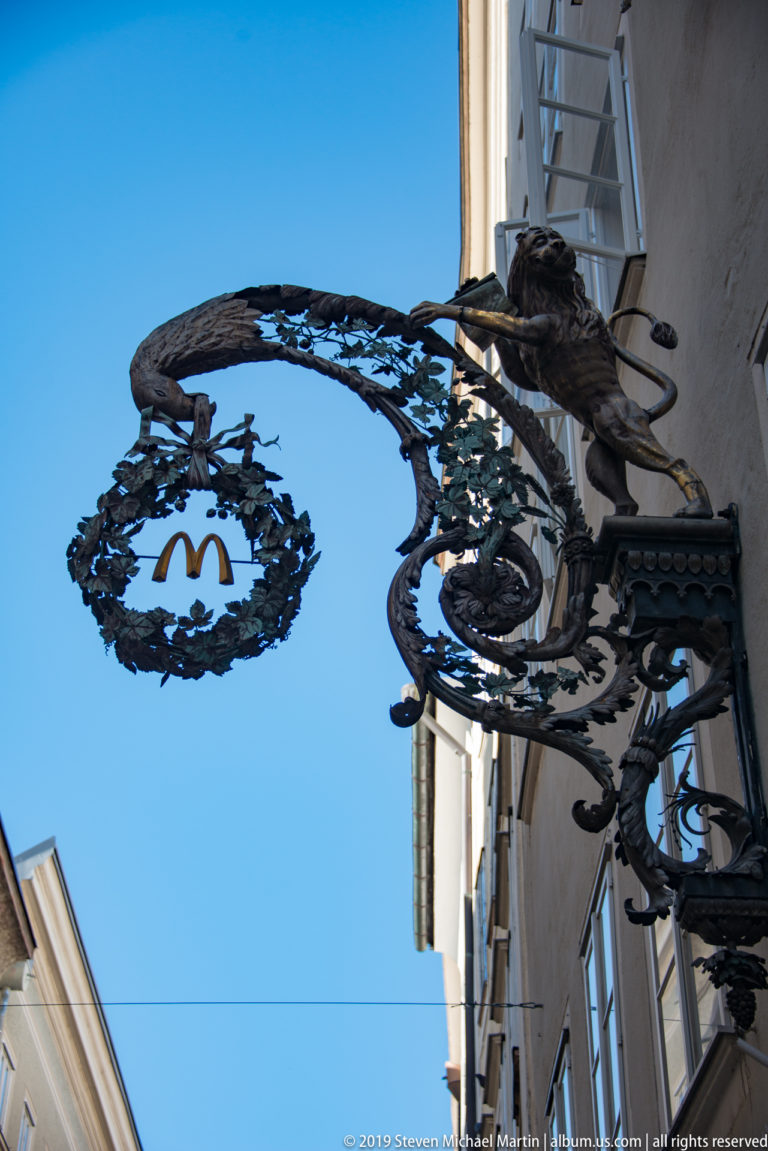 Low-angle view of an ornate black wrought-iron shop sign with a gilded emblem in a leafy circular wreath.