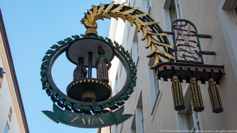 Circular green-and-gold wrought-iron ZARA sign with two stylized figures, mounted among other projecting shop signs.