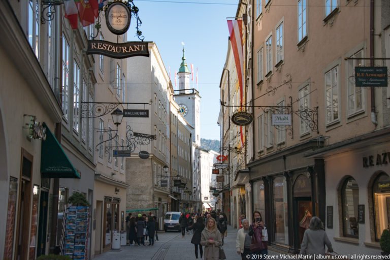 Pedestrians on narrow Getreidegasse beneath ornate hanging signs, with pale façades and a green-capped tower in the distance.